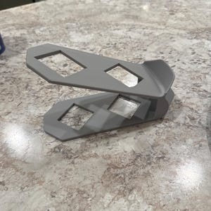 May include: A gray, 3D-printed phone stand with two angled platforms. Each platform has two square cutouts. The stand is designed to hold a phone in a vertical position. The stand is made of a smooth, matte material.