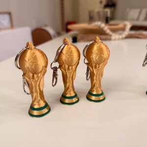 May include: Gold-colored trophy keychains with a green and gold base. The keychains are shaped like the FIFA World Cup trophy. The keychains have a silver keyring and clasp.