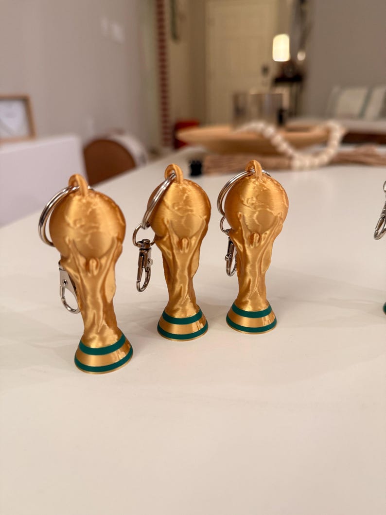 May include: Gold-colored trophy keychains with a green and gold base. The keychains are shaped like the FIFA World Cup trophy. The keychains have a silver keyring and clasp.