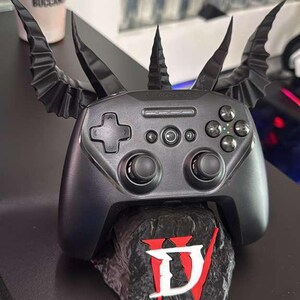 Puede incluir: Un mando de juegos negro con un dise&ntilde;o personalizado con cuernos y alas. El mando descansa sobre una base de piedra con el logotipo de Diablo IV en rojo y blanco. Un accesorio &uacute;nico para jugadores.