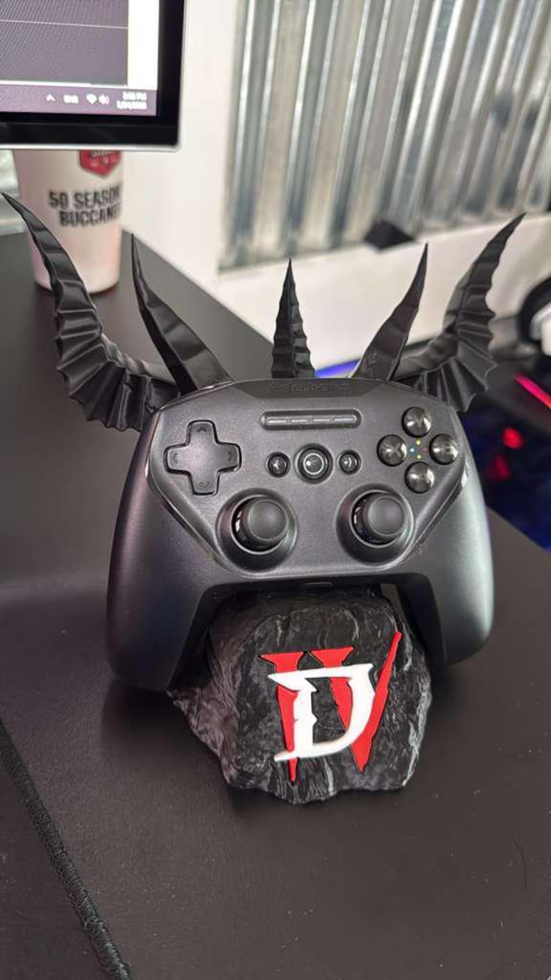 Puede incluir: Un mando de juegos negro con un dise&ntilde;o personalizado con cuernos y alas. El mando descansa sobre una base de piedra con el logotipo de Diablo IV en rojo y blanco. Un accesorio &uacute;nico para jugadores.