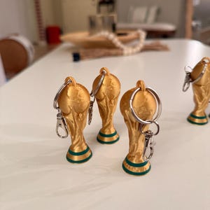 May include: Gold-colored trophy keychains with green accents on the base. The keychains have a silver keyring and clasp. The trophies are arranged on a white surface, suggesting a sports or awards theme.
