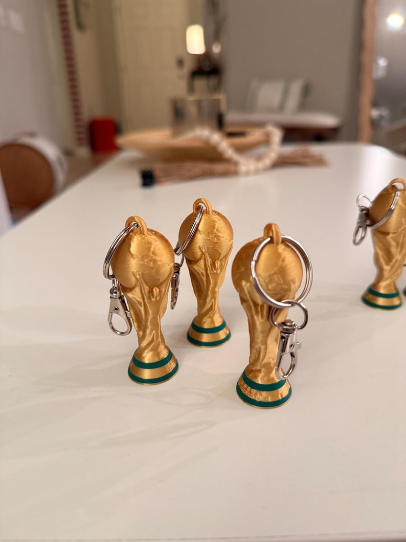 May include: Gold-colored trophy keychains with green accents on the base. The keychains have a silver keyring and clasp. The trophies are arranged on a white surface, suggesting a sports or awards theme.