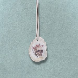 May include: An oyster shell-shaped ornament featuring a skull design with pink cherry blossoms. The ornament hangs from a cream ribbon. The skull is detailed with shading and the flowers are in full bloom. The background is a light teal colour.