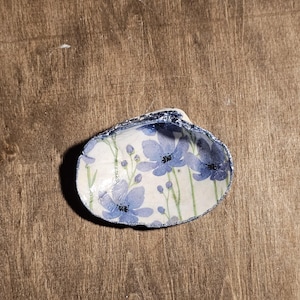 May include: A decorative seashell bowl with a floral design. The interior of the shell is painted with blue flowers and green stems on a white background. The rim of the shell is dark blue.