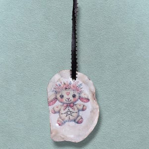 May include: A pendant with a bunny illustration, floral crown, and pentagram on a shell. The bunny has pink details and a bow. A black ribbon is attached for hanging.