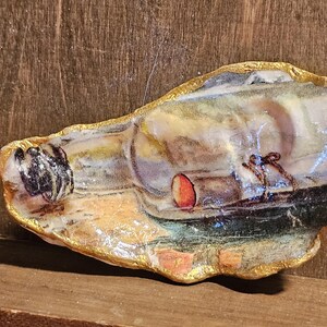 Peut inclure: Coquille d'huître peinte à la main avec une scène de paysage miniature et un bord doré. L'œuvre représente un bâtiment, un tonneau et des personnages, avec une palette de couleurs chaudes. La coquille est exposée sur une surface en bois.