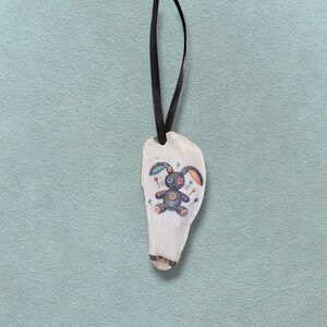 May include: A white, teardrop-shaped pendant with a black ribbon. The pendant features a colourful illustration of a bunny voodoo doll with button eyes and pins. The bunny has blue, orange, and black accents.