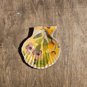 May include: A painted seashell with a floral pattern. The shell is yellow with pink, green, and orange floral details. It is placed on a wooden surface.