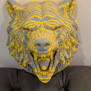 Può includere: Scultura tridimensionale a forma di testa di lupo con strisce gialle e grigie, che creano un effetto strutturato. La bocca del lupo è aperta, mostrando denti e lingua. La scultura è montata a parete.