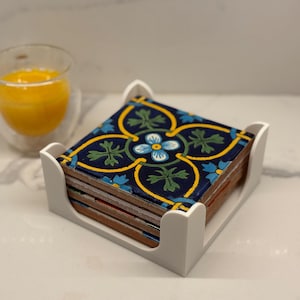 May include: A stack of square coasters with a blue, yellow, and green floral design, held in a white holder. A glass of orange juice sits in the background. Each coaster measures approximately 4 inches square.