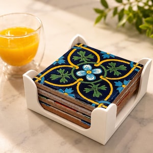 Könnte beinhalten: Ein Stapel quadratischer Keramikuntersetzer mit blauem, gelbem und grünem Blumendesign, in einem weißen Halter. Ein Glas Orangensaft steht neben den Untersetzern. Die Untersetzer sind etwa 10 cm groß.