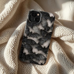 May include: A smartphone case featuring a repeating pattern of bats in shades of gray and white with gold accents. The case is shown resting on a cream-colored knitted blanket, showcasing the phone's design.