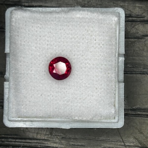 May include: A round, faceted, deep red gemstone, likely a ruby, rests in a white foam-lined plastic case. The case is square with a clear lid, set against a dark, textured wooden surface.