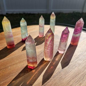 Puede incluir: Siete puntas de cristal de fluorita multicolores están dispuestas sobre una superficie de madera. Los cristales muestran una gama de colores, incluyendo rosa, púrpura, amarillo y verde. Cada cristal mide unos 10 cm de alto y tiene una punta.