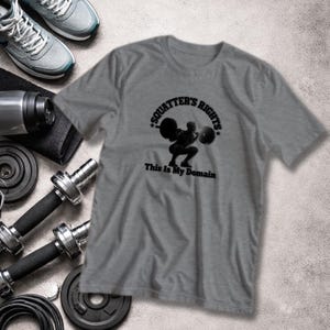 Op de afbeelding: Grijs t-shirt met de tekst "SQUATTER'S RIGHTS" en "This Is My Domain" in zwart. Het shirt heeft een afbeelding van een persoon die gewichten heft. Fitnessapparatuur, waaronder dumbbells, gewichten en sneakers, zijn zichtbaar.