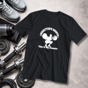 Op de afbeelding: Zwart t-shirt met de tekst "SQUATTER'S RIGHTS" en "This Is My Domain" in het wit. Het shirt heeft een afbeelding van een gewichtheffer. Fitnessapparatuur, waaronder dumbbells, gewichten en sportschoenen, is zichtbaar op de achtergrond.