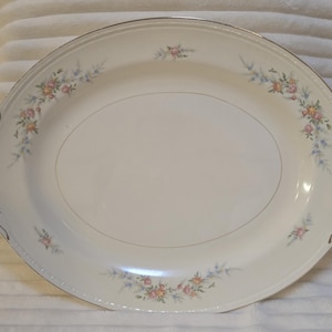 Könnte beinhalten: Eine ovale, cremefarbene Servierplatte mit goldenem Rand und floralen Akzenten. Die Platte hat zarte Ansammlungen von rosa, gelben und blauen Blumen. Die Griffe haben ein dekoratives Design.
