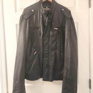 May include: Black leather jacket with a stand-up collar, shoulder accents, and multiple zippered pockets. The jacket is hanging on a wooden hanger against a white door. The jacket has a zipper closure and a small logo on the collar.