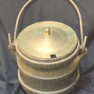 Puede incluir: Olla de cocina antigua de color bronce con tapa y asa. La olla tiene un cuerpo redondo con estrías horizontales y una tapa con un pequeño pomo. El asa es arqueada.