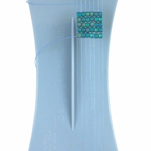 May include: A light blue Easy Jewel Loom with a partially completed beadwork square. The loom has a curved hourglass shape with vertical threads and a central guide. The beadwork features iridescent teal beads.