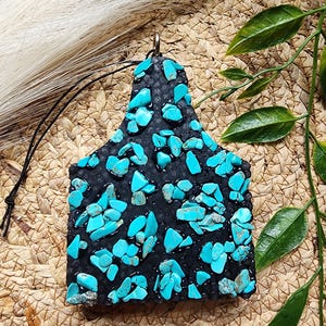 May include: A black pendant with turquoise stone accents, suspended by a black cord. The pendant has a tag shape and is set against a woven background with green leaves.