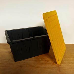 May include: A black rectangular storage bin with a yellow lid. The bin has a textured surface and is sitting on a wooden surface. The lid is leaning against the bin.