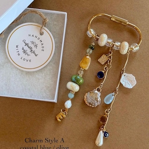 May include: A gold-toned oval keychain featuring a collection of dangling charms including a white shell, a blue teardrop, a small evil eye bead, and a pearl. A separate strand displays olive, yellow, and white beads with a gold letter E charm.
