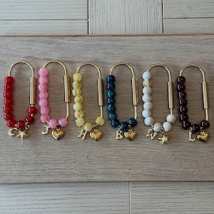 Può includere: Una collezione di sei portachiavi con elementi metallici color oro. Ogni portachiavi presenta perline di diversi colori, tra cui rosso, rosa, giallo, blu, bianco e marrone. Ogni portachiavi ha un ciondolo a forma di cuore e stella dorati.