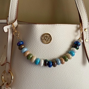 Puede incluir: Una correa de bolso con cuentas en tonos azules, verdes, marrones y crema, sujeta a un bolso blanco con herrajes dorados. La correa está hecha de cuentas redondas y se ve el texto "Coastal Earth".
