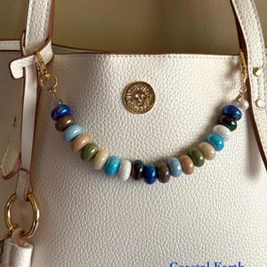 May include: A white textured tote bag featuring a gold lion emblem and a decorative beaded chain attached to the handles. The chain consists of smooth, round, multi-coloured beads in shades of blue, green, brown, and white. The text Coastal Earth is visible.