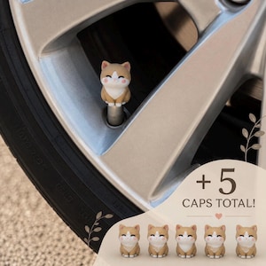 May include: A set of five tyre valve caps, each featuring a small, tan and white cat figurine with pink cheeks. One cap is installed on a silver car wheel valve stem, while the other four are displayed below with the text + 5 CAPS TOTAL!