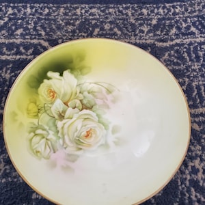 Puede incluir: Un cuenco redondo de porcelana decorativo con un diseño floral pintado a mano. El cuenco presenta rosas blancas con acentos amarillos y verdes, sobre un fondo amarillo pálido y blanco. El borde tiene un ribete dorado.