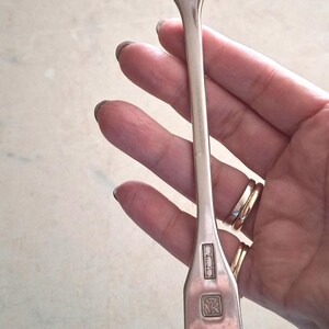 May include: A silver spoon with a rounded bowl and a long, rectangular handle. The handle has a hallmark with the initials "MK" and other markings. The spoon is held in a hand.