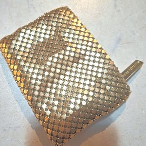 Könnte beinhalten: Eine Vintage-Geldbörse aus goldfarbenem Mesh mit rechteckiger Form. Die Geldbörse besteht aus kleinen, kreisförmigen, ineinandergreifenden Metallteilen. Ein kleiner, rechteckiger Verschluss ist an der Seite befestigt. Die Geldbörse hat eine glänzende Goldfarbe.