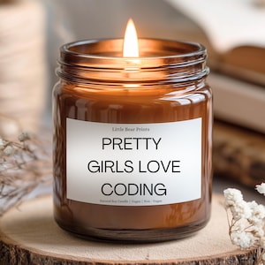 Peut inclure: Une bougie en pot de verre brun allumée avec le texte "PRETTY GIRLS LOVE CODING" sur une étiquette blanche. La bougie est sur une surface en bois avec de petites fleurs blanches. La bougie est une bougie de soja naturelle et est végétalienne.