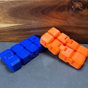 May include: Two fidget toys are shown, one blue and one orange. Each toy is made up of connected, rectangular blocks. The blue toy is on the left, and the orange toy is on the right. The background is a wooden surface and a grey surface.