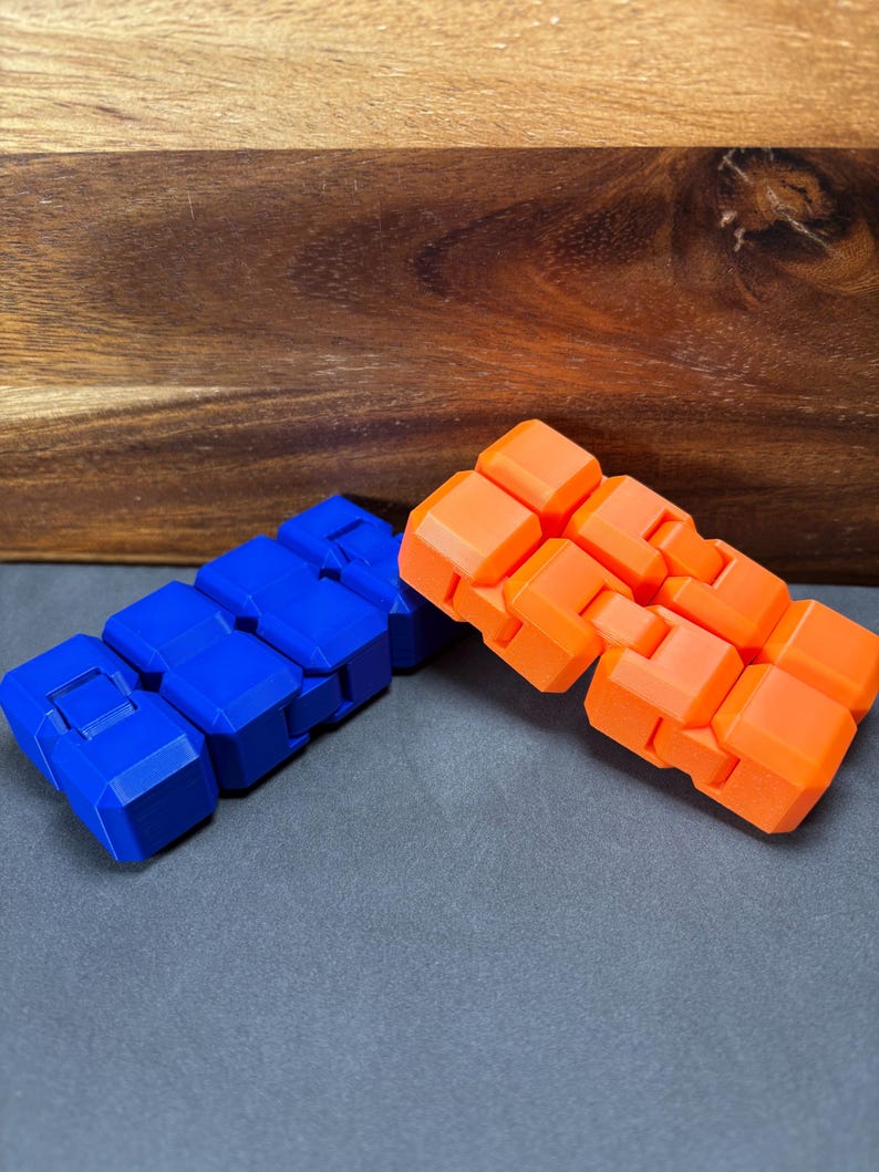 May include: Two fidget toys are shown, one blue and one orange. Each toy is made up of connected, rectangular blocks. The blue toy is on the left, and the orange toy is on the right. The background is a wooden surface and a grey surface.