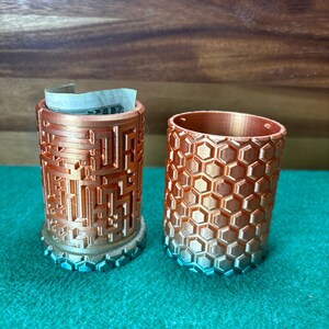 May include: Two cylindrical containers with a metallic gradient finish. One has a geometric pattern, and the other has a honeycomb design. A bill is partially visible inside the geometric container. The containers are on a green surface.