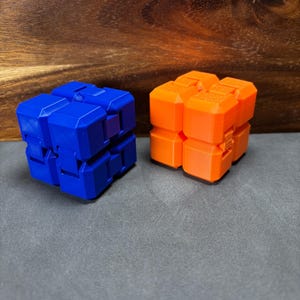 May include: Two geometric fidget toys. One is blue, the other is orange. Each is composed of multiple connected cube-shaped segments. The toys are on a grey surface with a wooden background.