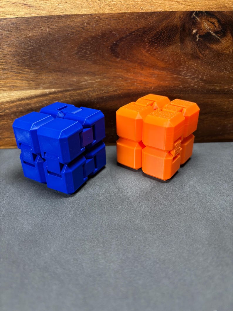 May include: Two geometric fidget toys. One is blue, the other is orange. Each is composed of multiple connected cube-shaped segments. The toys are on a grey surface with a wooden background.
