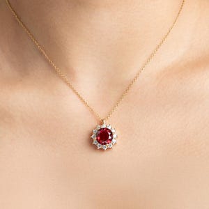 Op de afbeelding: Een gouden ketting met een hanger met een grote, ronde, dieprode edelsteen omringd door kleinere, heldere edelstenen. De hanger hangt aan een fijne gouden ketting, een klassiek en elegant sieraad.