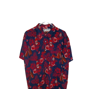 May include: Short-sleeved button-down shirt with a navy blue background and a pattern of red hibiscus flowers, bottles, and mugs. The shirt has a collar and a few visible buttons. The text "KAMS" is visible on the shirt.