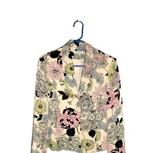 May include: A cream-colored blazer with a vibrant floral pattern in pink, black, yellow, and blue. The jacket is hung on a blue hanger. The floral print covers the entire jacket.