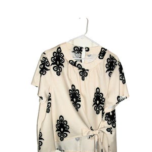 May include: A short-sleeved cream-colored blouse featuring a repeating black damask-style pattern. The garment has a round neckline with a small back button closure and a side tie detail at the waist that creates a wrap-effect silhouette.