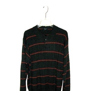 May include: A dark gray sweater with red horizontal stripes. The long-sleeved sweater has a ribbed texture and a button-up collar. The sweater is hanging on a clear plastic hanger against a white background.