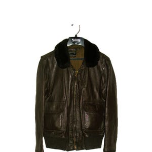 May include: A brown leather jacket with a black fur collar, two front pockets, and a zipper closure. The jacket is hanging on a black hanger. The jacket has a ribbed hem and cuffs.