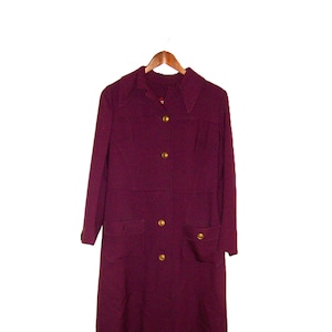 May include: A long, burgundy dress coat with a collared neckline and gold-colored buttons. The coat features two front pockets and is hanging on a wooden hanger. The coat is a solid color and appears to be made of a woven fabric.