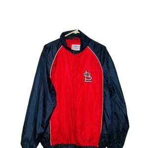 May include: A red and navy blue windbreaker jacket with a white trim. The jacket features the St. Louis Cardinals logo on the front. The jacket is hanging on a white hanger.