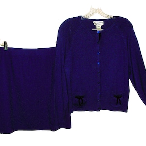 May include: A purple cardigan with a button fastening and black bow details on the pockets. The cardigan is paired with a matching purple skirt.